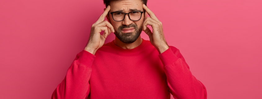 Troubled distressed bearded man keeps index fingers on temples to focuse, tries to concentrate on task, frowns and grimaces, has huge headache or migraine, bothered with painful disease, models indoor